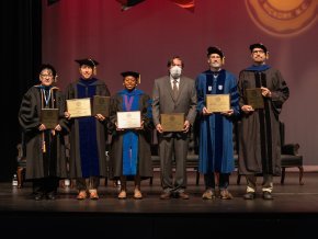 Six faculty members in full regalia stand on an indoor stage and display their awards