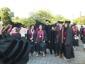 Five students stand in front of a professor with arms spread wide, all wearing commencement regalia