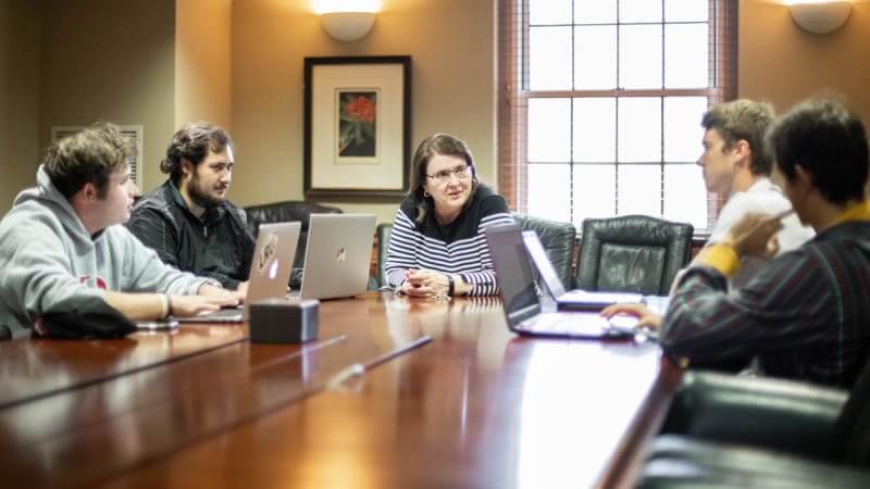 Investing club students sitting around conference table discuss recent investments with professor