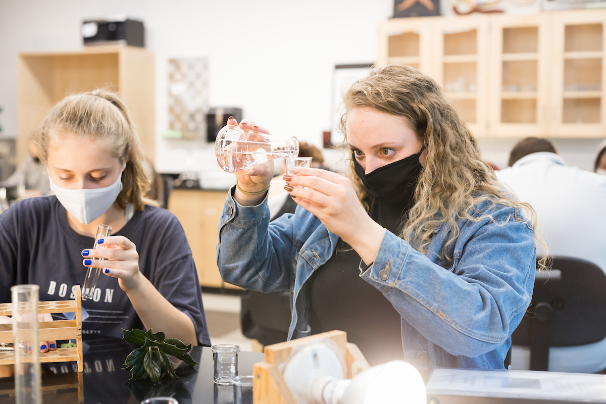 Amy Sain, right, pours liquid from a beaker while masked in a lab