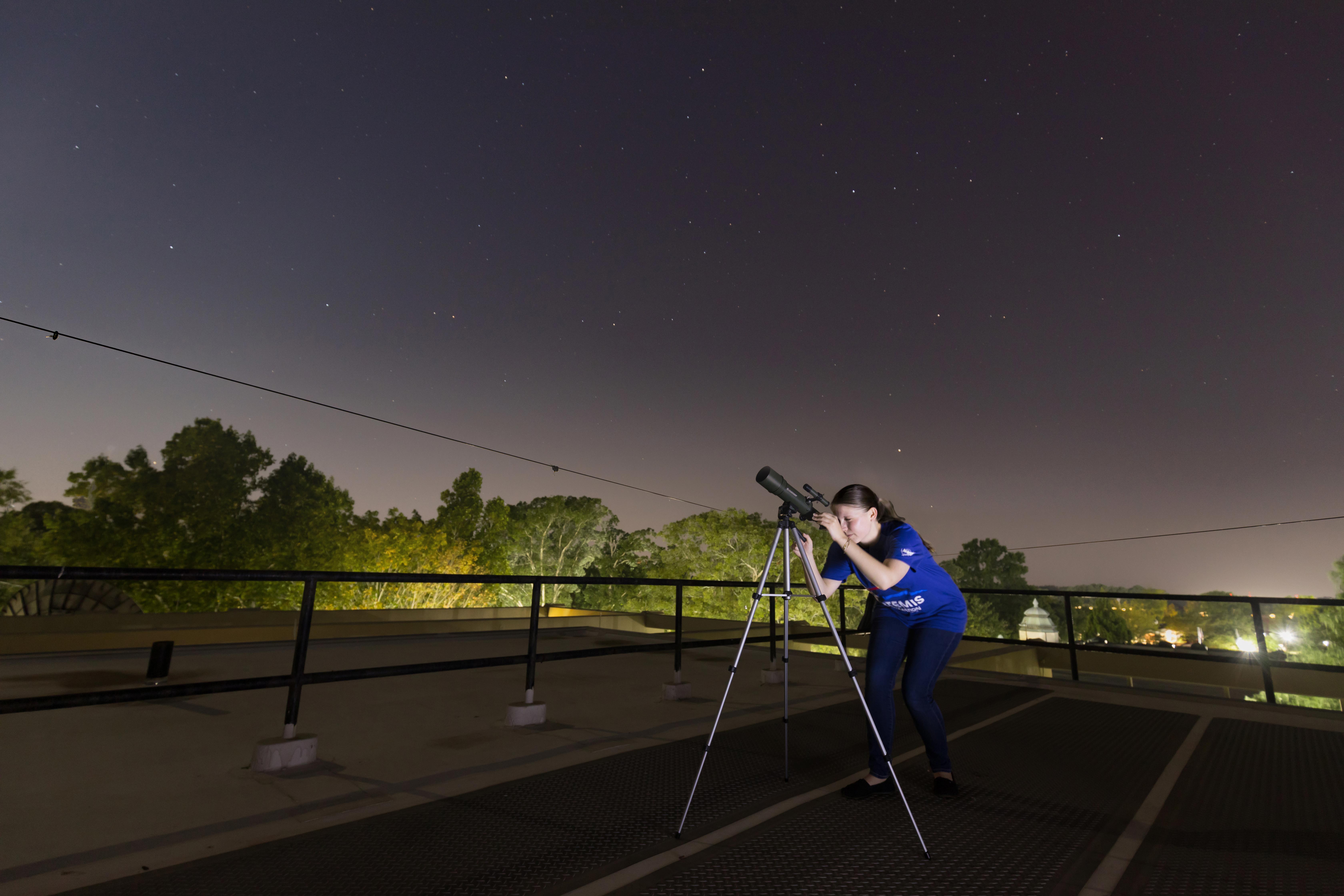 Madeline Schmidt stands outside and looks through a telescope at night.