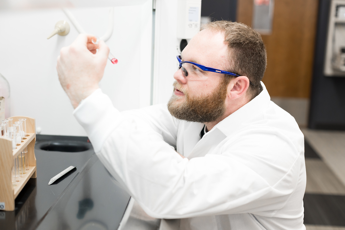 Noah Jenkins wears a lab coat and safety glasses in an indoor lab