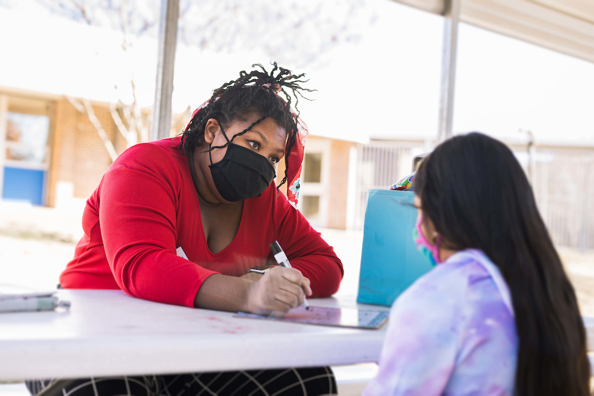 While wearing a mask, Zakiya Ruth, left, writes on a whiteboard on the table while looking at a student who has her back to the camera.
