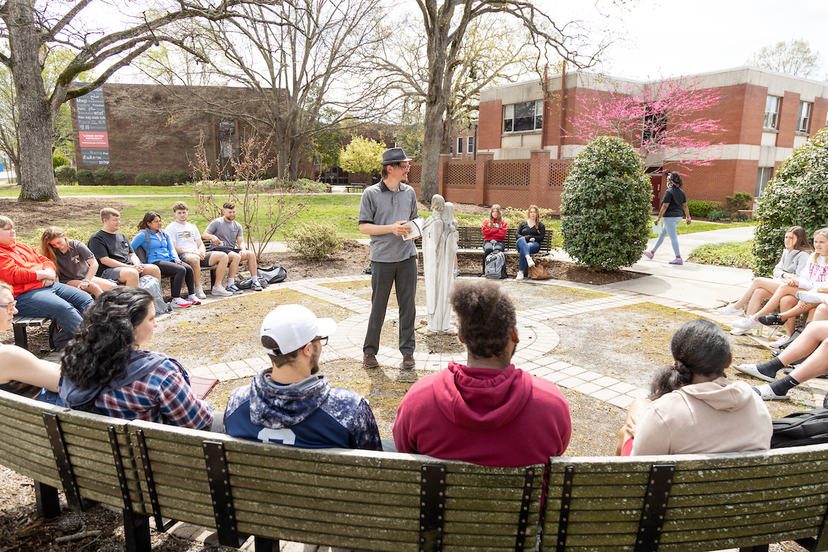 A class sits on benches outside as a professor stands and speaks to them