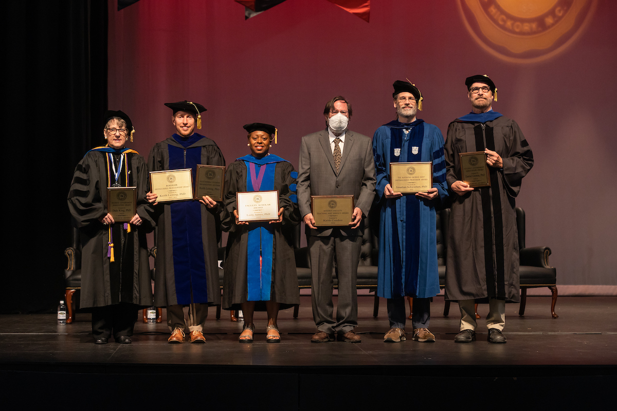 Six faculty members in full regalia stand on an indoor stage and display their awards