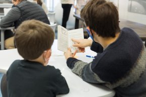 A teacher and student look at a book while sitting indoors with their back to the camera