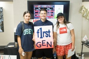 Three students stand together and smile at the camera while holding a sign