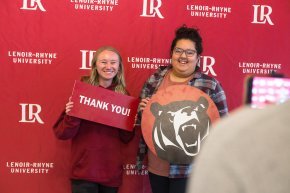 Two students hold up signs against a red and white backdrop