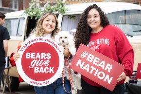 Two female students smile while holding signs and a little dog while outside