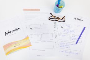 A journal, papers, a pen, cup and glasses sit on a white desk