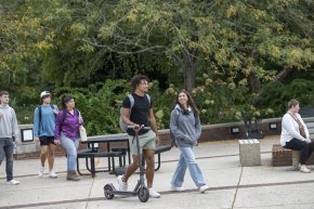 One student wheels a scooter across Shaw Plaza while surrounded by other students ahead and behind