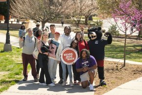 Nine students pose with Joe Bear holding Bears Give Back signs with trees and the quad in the backgroun