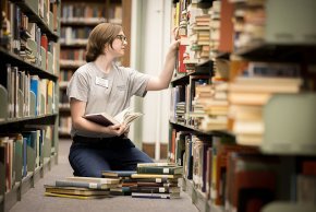 Lucas Quinn sits on the floor between stacks in the library and shelves a small pile of books