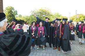 Five students stand in front of a professor with arms spread wide, all wearing commencement regalia