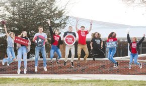 9 students holding thank you signs jump in the air in front of Shaw Plaza