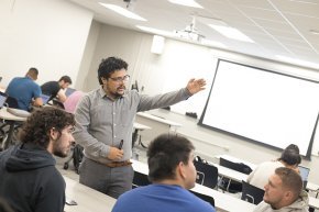 A professor gestures toward the projection screen in a full classroom