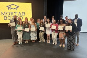 A dozen or so current Mortar Board members display their certificates in front of a lighted gold Mortar Board logo