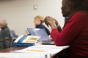 Close up of a woman at a desk in a classroom, hands poised over a textbook on Christianity