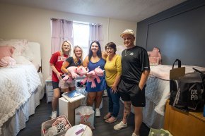 Two roommates holding matching pink rhinoceros stuffies pose in a dorm room with their parents