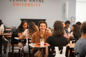 Student smiles across the table at other students in the dining hall