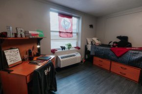 View of model dorm room with desk left, window center and bed right
