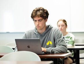 A student works at his laptop in Creative Collaboration and Culture. A second student works at the desk behind him