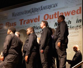 Tenor and baritone sections of the choir on stage in front of a projection of a newspaper page from the Freedom Rides