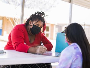 While wearing a mask, Zakiya Ruth, left, writes on a whiteboard on the table while looking at a student who has her back to the camera.