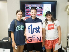 Three students stand together and smile at the camera while holding a sign