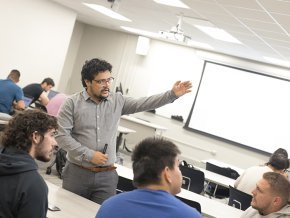 A professor gestures toward the projection screen in a full classroom