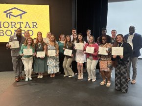 A dozen or so current Mortar Board members display their certificates in front of a lighted gold Mortar Board logo 