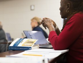 Close up of a woman at a desk in a classroom, hands poised over a textbook on Christianity