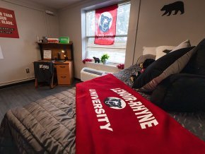 Model dorm room with bed and red LR blanket in close foreground, window and desk further back