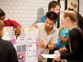 Students explain research findings to their peers over a display on SOURCE Day