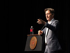 Andrew McCarthy leans on a podium with the LR seal on front