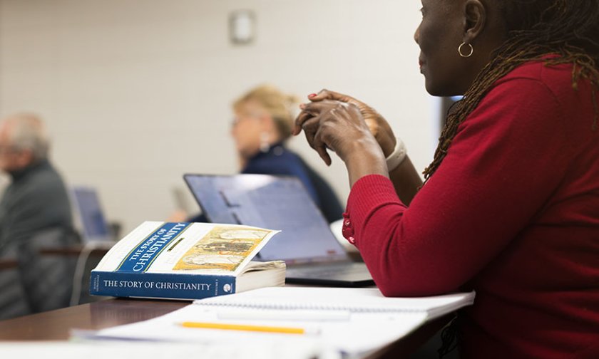 Close up of a woman at a desk in a classroom, hands poised over a textbook on Christianity