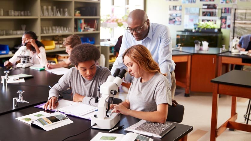 Science teacher overlooks shoulder of student working with microscope