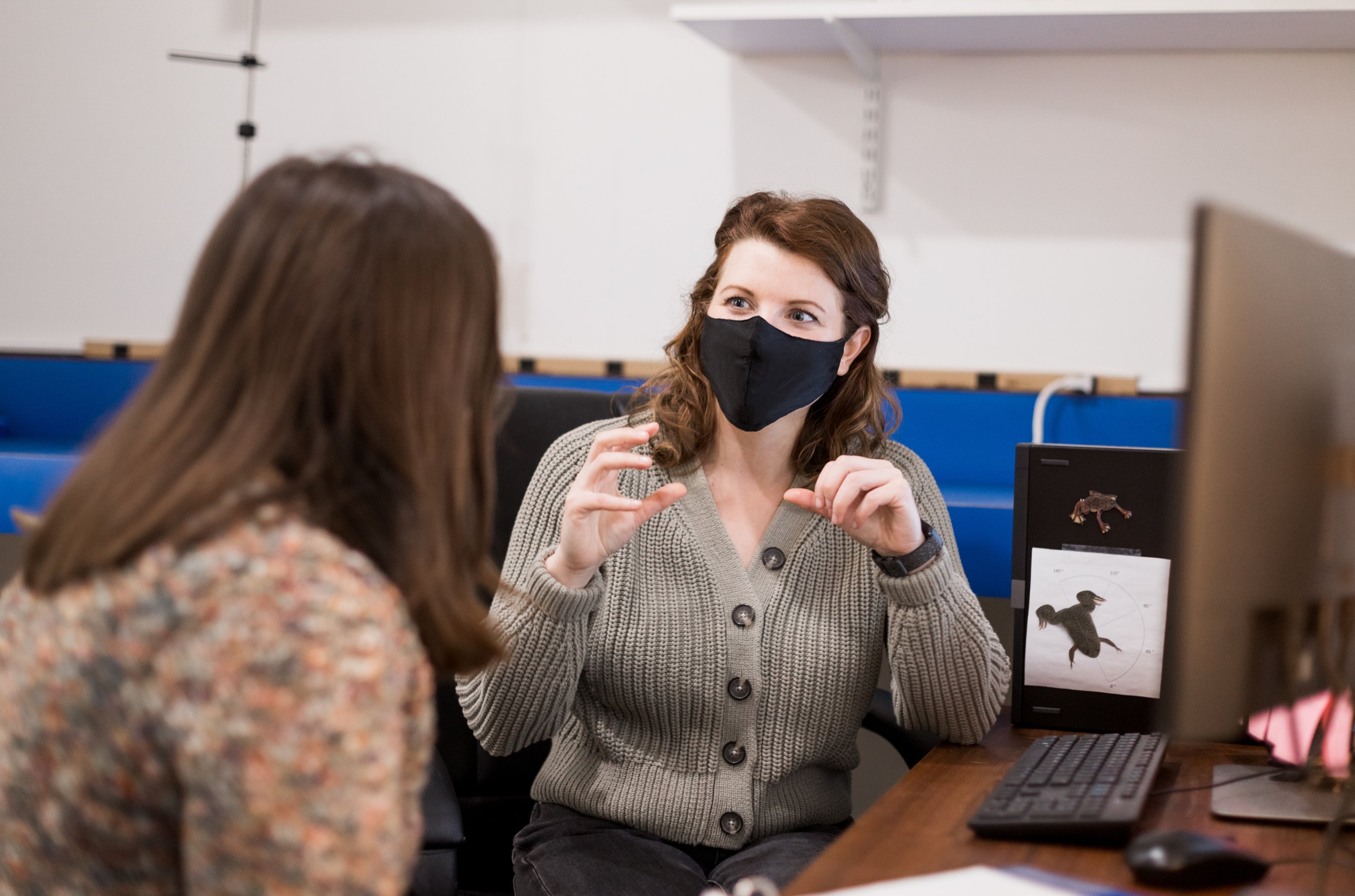 Maggie Keller, with her back to the camera, talks to Carly York, masked, in her office