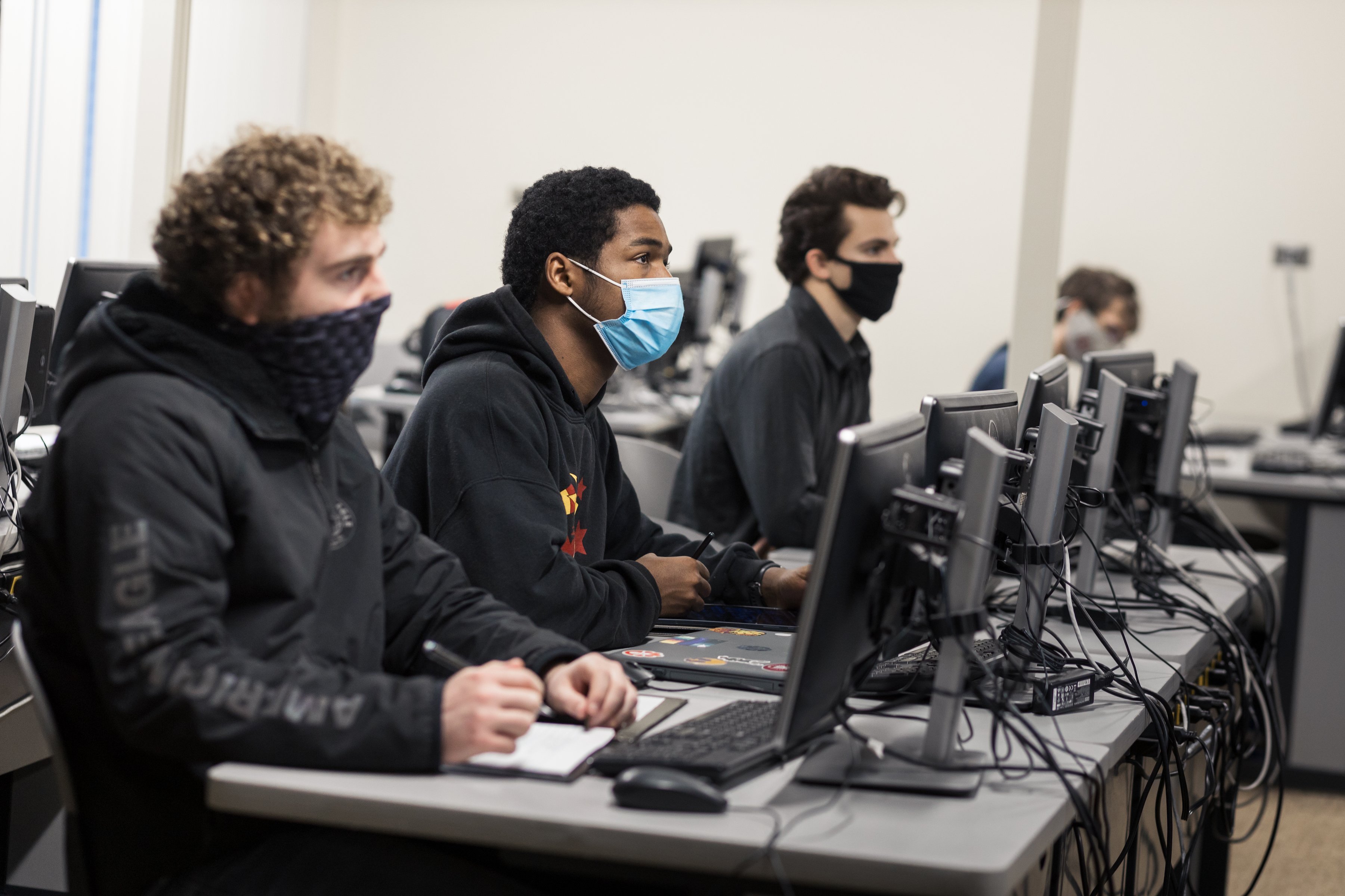 Students sit in a classroom in front of computers while wearing masks