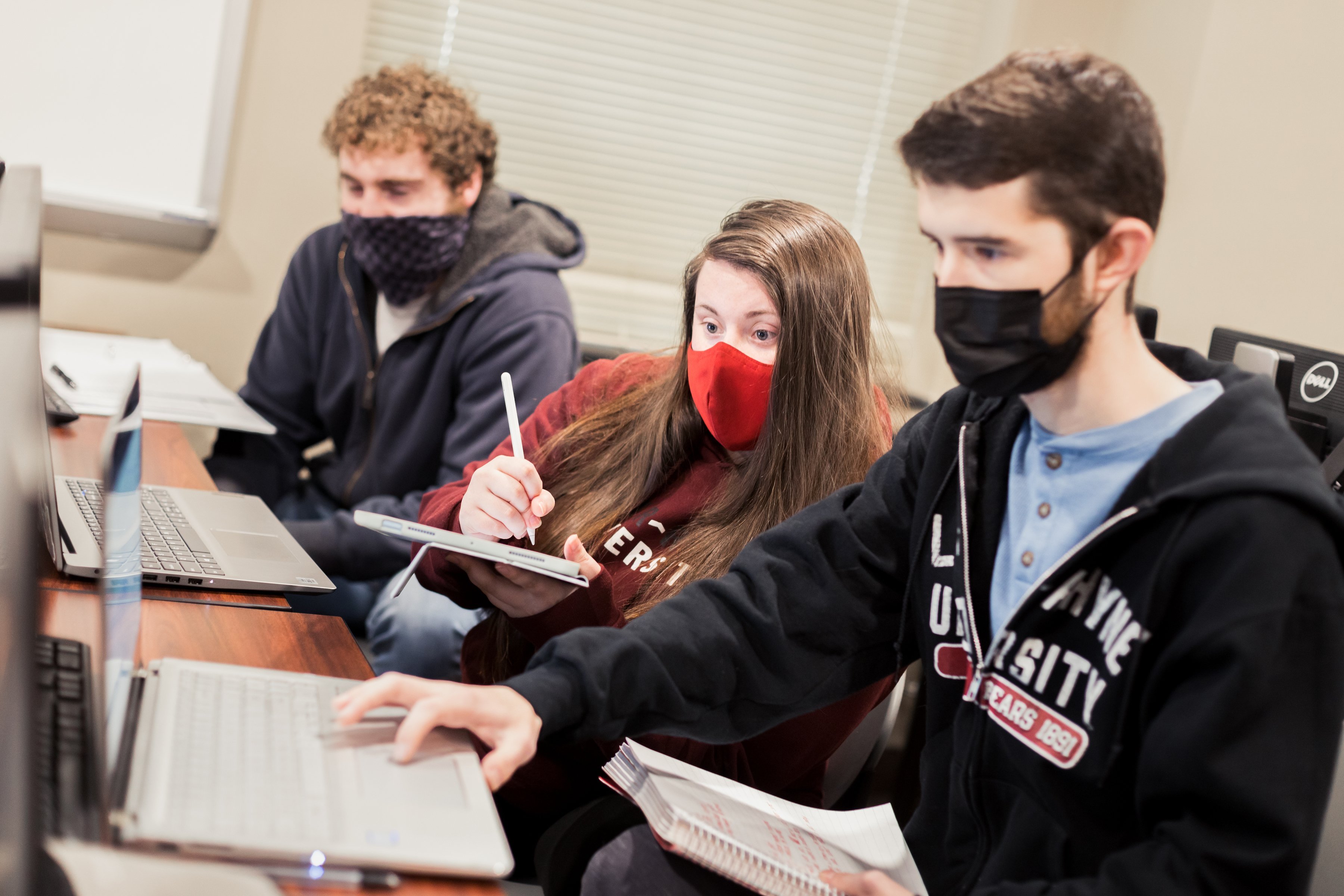 Three masked students sit in a classroom taking notes and working on computers