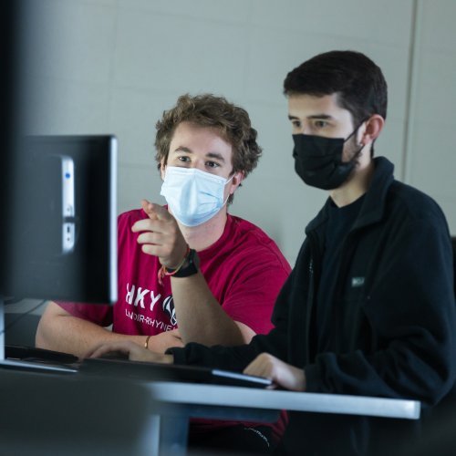 Two masked students sit in a classroom and look at the same computer screen