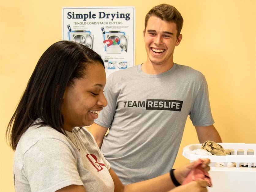 Two students smile while folding laundry in a dorm laundry room.