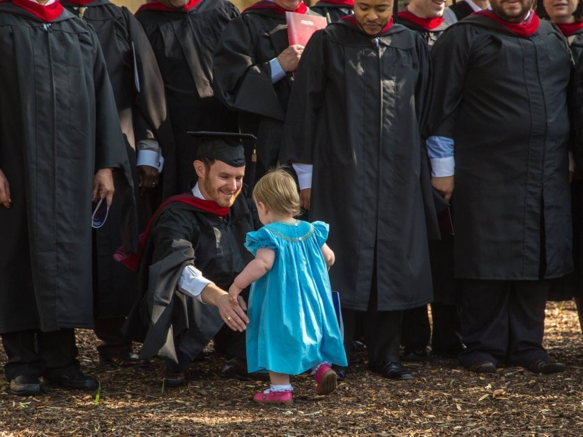 A graduate kneels down to hug his young daughter.