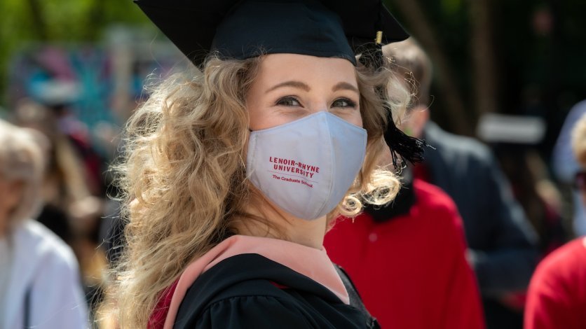 A graduate student looks back the camera and smiles while wearing a mask outdoors.